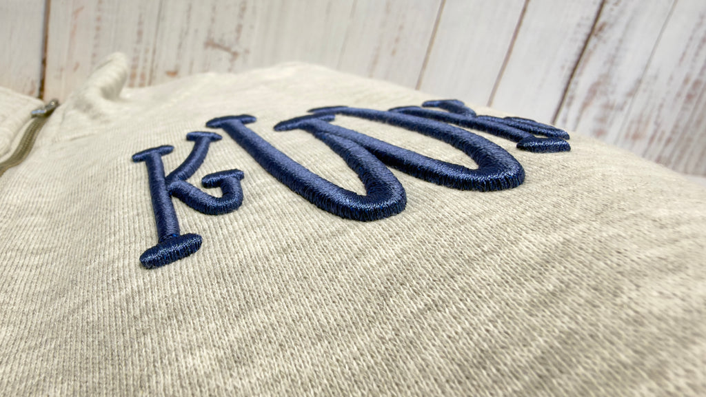 Grey quarter zip sweatshirt with blue 3D puff embroidered letters on a wooden surface