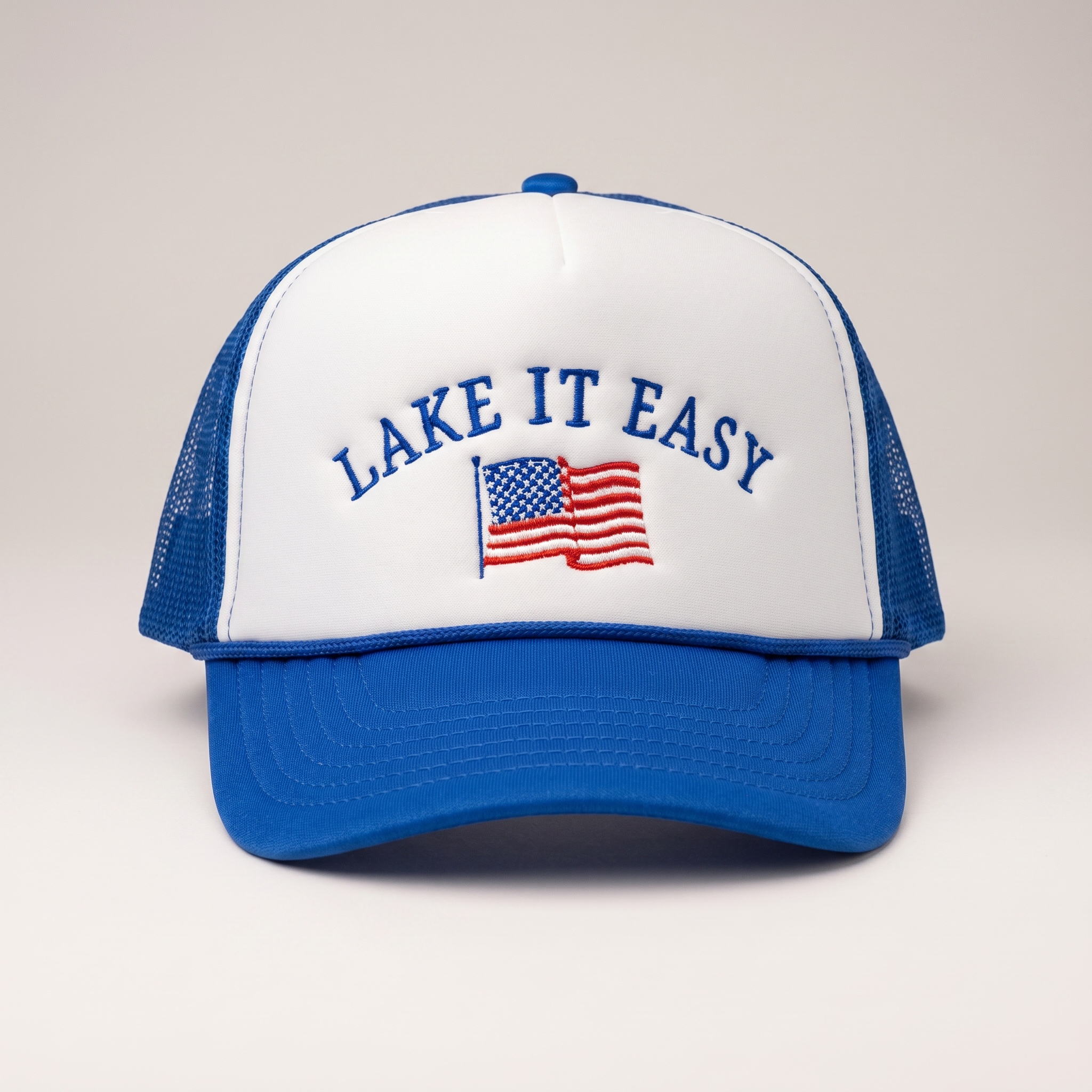 Blue and white cap with 'Lake It Easy' text and American flag on a light gray background.  Foam trucker cap with rope.