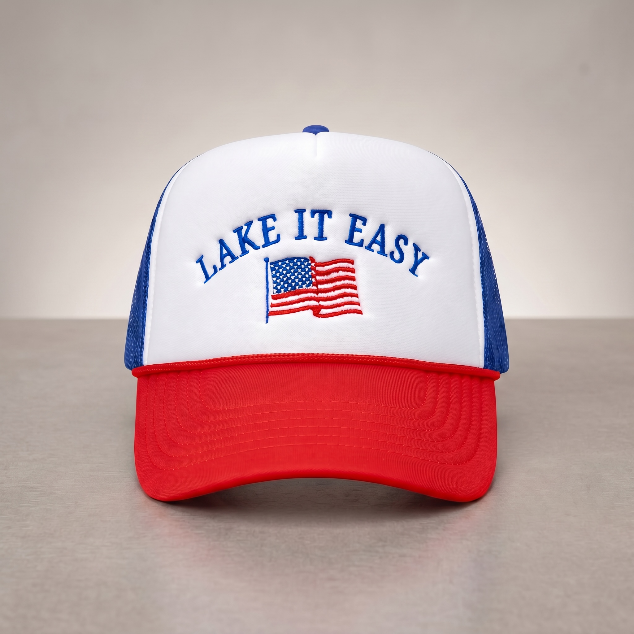 Trucker hat with 'Lake It Easy' text and American flag on a neutral background.  Foam trucker with rope embroidered cap.