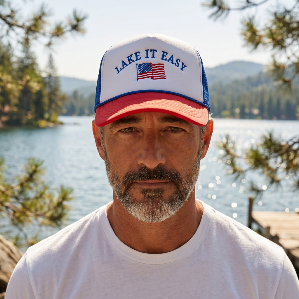 Man wearing a 'Lake It Easy' cap with an American flag, standing by a lake with trees and mountains in the background.
