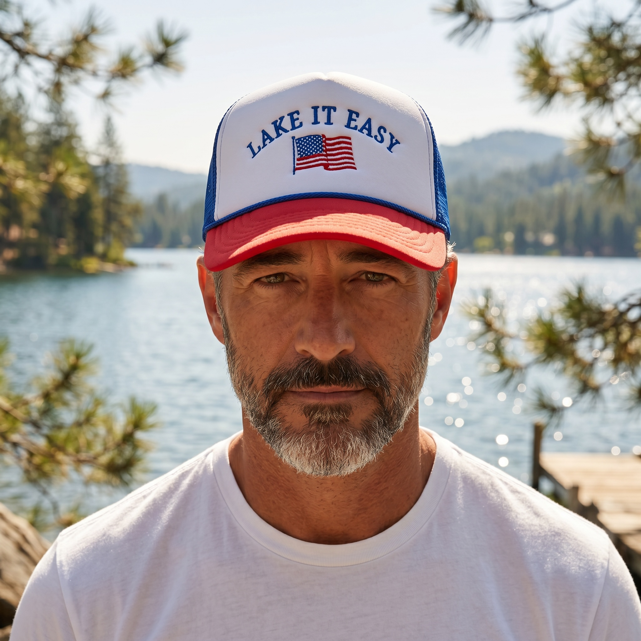 Man wearing a 'Lake It Easy' cap with an American flag, standing by a lake with trees and mountains in the background.
