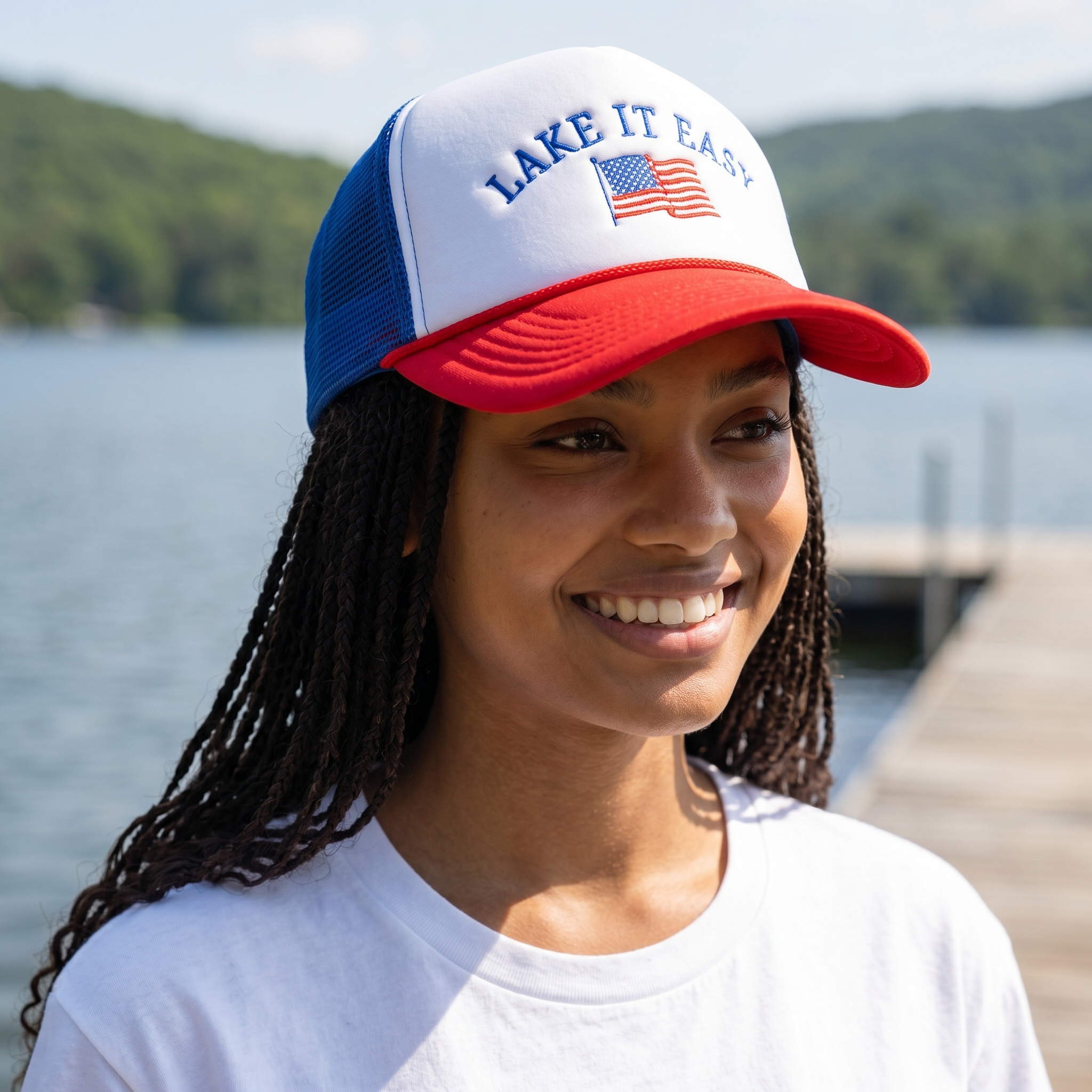 Woman wearing a foam rope trucker cap 'Lake It Easy' embroidered cap with an American flag, standing by a lake.