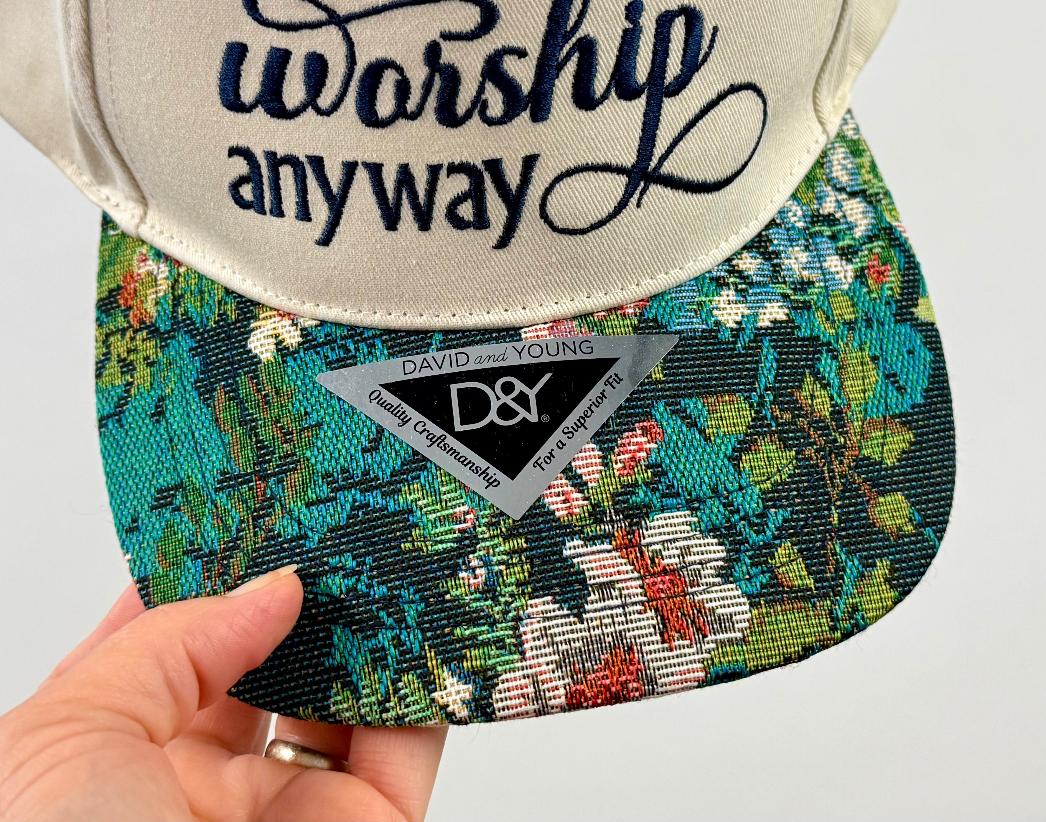 Cap with 'worship anyway' text and floral pattern, held by a hand.